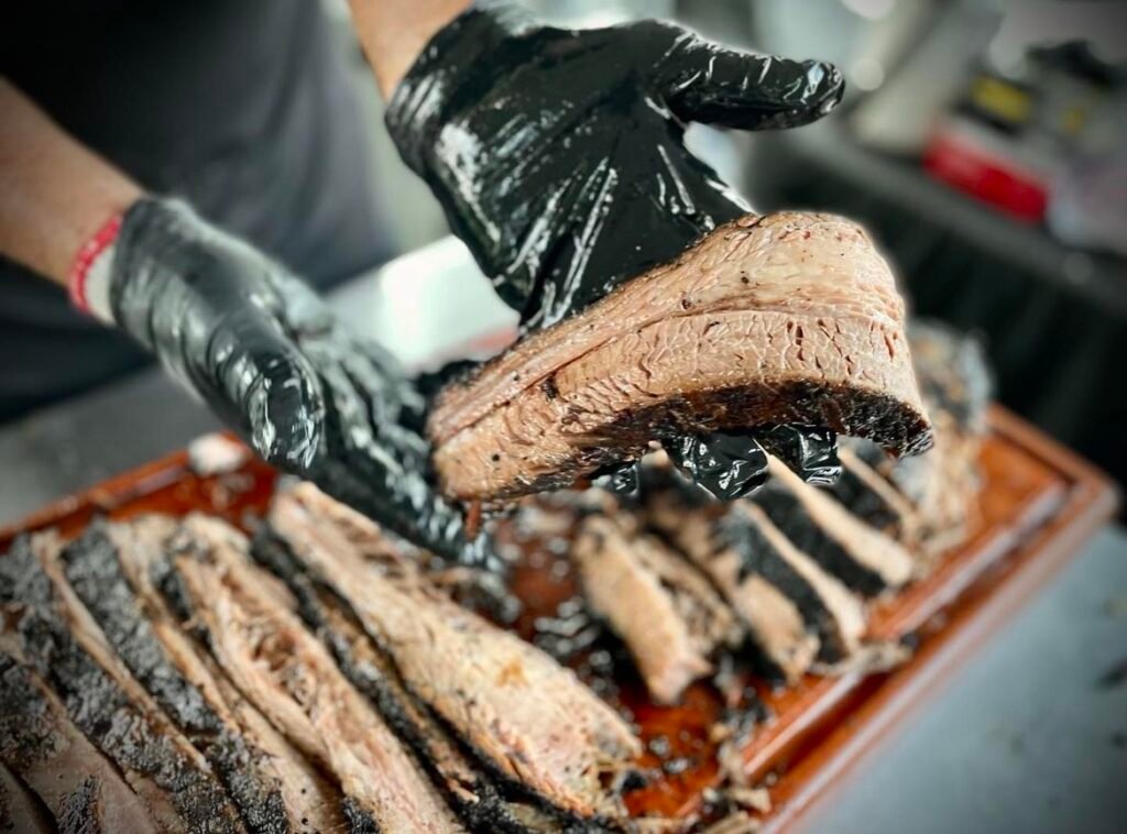 A person wearing black gloves holding a slice of beef brisket on top of a cutting board of beef brisket.